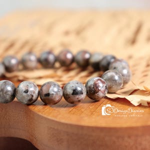 May include: A beaded bracelet featuring round, gray and brown speckled stones. The bracelet is displayed on a wooden surface with a light brown paper background. The image includes the text "SJ Design Diaries Healing Crystals".