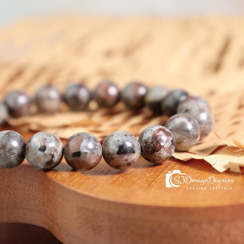 May include: A beaded bracelet featuring round, gray and brown speckled stones. The bracelet is displayed on a wooden surface with a light brown paper background. The image includes the text "SJ Design Diaries Healing Crystals".