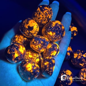 Puede incluir: Un conjunto de piedras de sodalita brillantes, sostenidas en una mano. Las piedras tienen un patrón moteado de naranja y negro, con un brillo fluorescente brillante. El fondo oscuro resalta los colores vibrantes de las piedras.