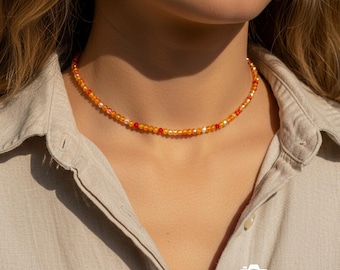 May include: A beaded necklace with orange, red, and white beads. The necklace is worn around the neck and is made of small, round beads. The necklace is a choker style and is likely handmade. The necklace is a piece of jewelry.