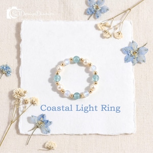 May include: A beaded ring featuring alternating light blue and white spherical beads interspersed with small gold-toned metallic spheres. The text Coastal Light Ring is printed below the ring on a square piece of textured white paper.