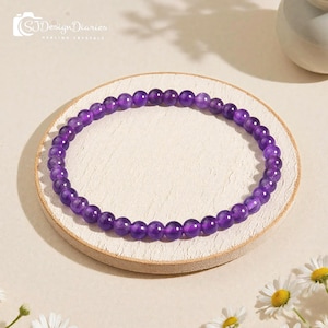 May include: A purple amethyst beaded bracelet. The bracelet is made of small, round, polished amethyst beads strung together. The bracelet is resting on a small, round, off-white wooden tray. The background is a light beige color with a few daisies.