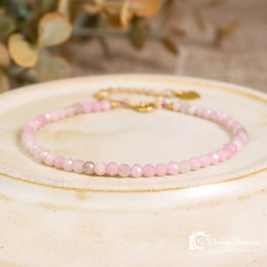 May include: A delicate pink beaded bracelet with a gold chain clasp. The bracelet is made of small, faceted pink beads. The background is a neutral cream-colored surface with a blurred green plant.
