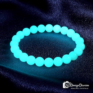 Puede incluir: Una pulsera circular compuesta por cuentas esféricas lisas que emiten un brillo cian brillante y luminoso. Las cuentas están dispuestas en un bucle continuo, creando un efecto de luz vibrante sobre la superficie oscura y texturizada de fondo.