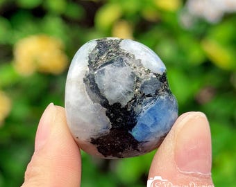 May include: A white, blue, and black gemstone with a smooth, rounded surface. The gemstone is being held by a hand.