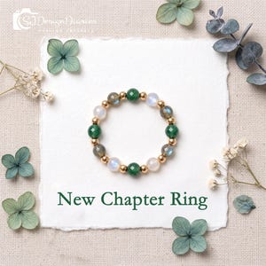 May include: A beaded ring featuring alternating round faceted green, grey, and translucent white beads interspersed with small gold-toned metallic spheres. The text New Chapter Ring is printed in dark green below the circular beaded ring.