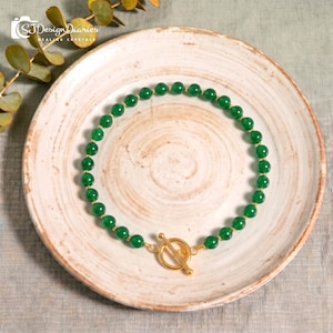Puede incluir: Un collar que presenta una hilera continua de cuentas redondas y lisas de color verde intenso, intercaladas con pequeños espaciadores de metal dorado. La pieza se asegura con un cierre de palanca de metal dorado formado por un anillo y una barra.