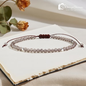 Peut inclure: Un bracelet de perles avec de petites perles grises à facettes enfilées sur un cordon marron foncé. Le bracelet a une fermeture coulissante et de petites perles argentées. Le fond comprend des fleurs séchées et le texte "SJ Design Diaries Healing Crystals."