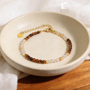 May include: A beaded bracelet featuring alternating sections of brown tiger eye and clear quartz gemstones. The bracelet has a gold-colored chain and clasp, with a small heart-shaped charm. The bracelet is displayed on a white ceramic dish.