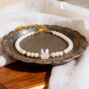 May include: A beaded bracelet featuring small white round beads and gold-toned metal spacers, centered with a white rabbit-shaped charm. The bracelet includes a gold-toned metal chain extension with a small heart-shaped charm at the end.