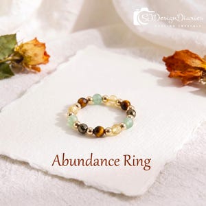 May include: A beaded ring featuring alternating round tiger eye, light green, and yellow faceted beads interspersed with small metallic gold-tone spheres. The text Abundance Ring is printed below the ring, and SJDesignDiaries Healing Crystals is at the top.