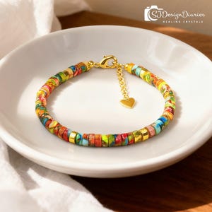 May include: A colorful beaded bracelet with a gold clasp and heart charm. The bracelet features a mix of red, blue, yellow, and green beads. The text "SJDesignDiaries Healing Crystals" is in the upper right corner.