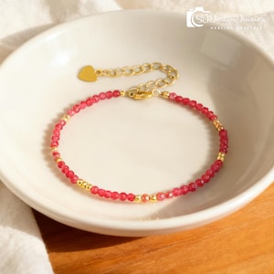 May include: A delicate bracelet featuring small, faceted red beads interspersed with gold-coloured beads. The bracelet has a gold-coloured chain and a heart-shaped charm. The bracelet is displayed on a white ceramic bowl. The text "SJ Design Diaries" is visible.