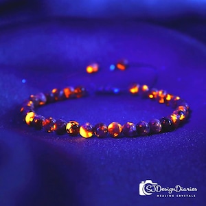 May include: A beaded bracelet featuring round, dark purple stones that exhibit a bright, glowing orange fluorescence under ultraviolet light. The bracelet is arranged in a circular shape with two additional loose beads visible in the background.