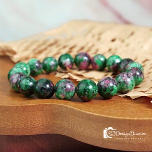 May include: A beaded bracelet featuring round, polished gemstones. The beads are primarily green with red and black inclusions. The bracelet is displayed on a wooden surface with a piece of textured paper in the background. The image includes the text "SJ Design Diaries Healing Crystals."