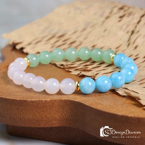 Pode incluir: Uma pulseira de contas com pedras preciosas rosa claro, verde claro e azul claro. A pulseira tem detalhes dourados. É exibida em uma superfície de madeira com um fundo de papel. O texto "SJDesignDiaries Healing Crystals" é visível.