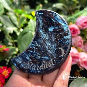 Op de afbeelding: Een maanvormig kristal met een blauw en zwart patroon en het woord "Stardust" gegraveerd. Een kleine zilveren maan is ook op het kristal gegraveerd. Het kristal wordt in een hand gehouden, met een achtergrond van roze bloemen en groen gebladerte.