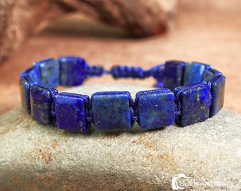 Pode incluir: Uma pulseira de lápis-lazúli azul com contas quadradas. A pulseira é enfiada em um cordão azul e repousa sobre uma pedra cinza. As contas de lápis-lazúli têm manchas douradas.