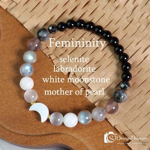 May include: A beaded bracelet featuring a gradient of gemstones including black beads, grey labradorite, iridescent moonstone, and white mother of pearl. The bracelet includes a white crescent moon charm and text reading Femininity, selenite, labradorite, white moonstone, mother of pearl.