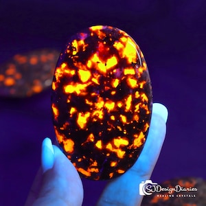 May include: An oval-shaped, polished stone with a striking pattern of bright orange and yellow inclusions against a dark background. The stone is held in a hand with a light blue manicure. The image is lit with a purple hue.