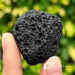 Könnte beinhalten: Ein dunkelgraues, poröses Vulkangestein, das zwischen Fingern gehalten wird. Der Stein hat eine raue, strukturierte Oberfläche mit zahlreichen kleinen und großen Löchern. Der Hintergrund ist verschwommenes grünes Laub. Der Text "SJ Design Diaries Healing Crystals" ist enthalten.