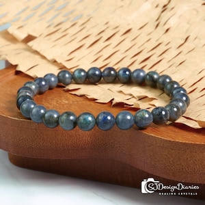 Op de afbeelding: Een kralenarmband met donkerblauwe en groene ronde kralen. De armband wordt gepresenteerd op een houten dienblad met een lichtbruine papieren achtergrond. De kralen hebben een gepolijst oppervlak. De armband is een sieraad. De kralen zijn ongeveer 0,8 cm in diameter.