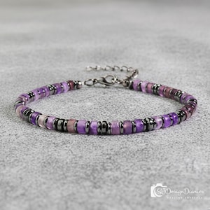May include: A beaded bracelet featuring a mix of purple and dark gray beads. The bracelet has a silver-colored clasp and is displayed on a gray surface. The beads are a mix of purple hues, with some dark gray accents.
