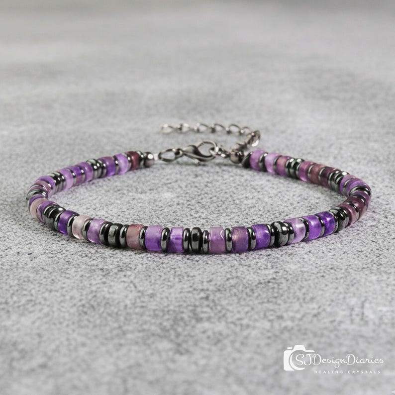 May include: A beaded bracelet featuring a mix of purple and dark gray beads. The bracelet has a silver-colored clasp and is displayed on a gray surface. The beads are a mix of purple hues, with some dark gray accents.