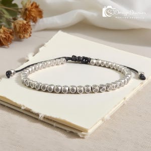 May include: Silver beaded bracelet with a black adjustable cord. The bracelet features small, geometric beads with a floral design. The bracelet is displayed on a stack of cream-colored paper.