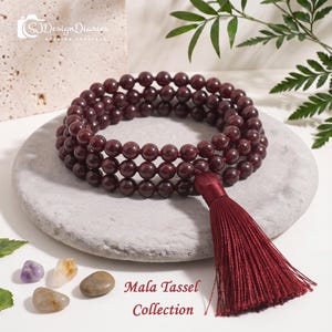May include: A multi-strand mala necklace featuring deep red, polished spherical beads and a matching dark red silk tassel. The text "SJDesignDiaries HEALING CRYSTALS" and "Mala Tassel Collection" is visible. Three small, smooth, multi-colored stones are placed nearby.