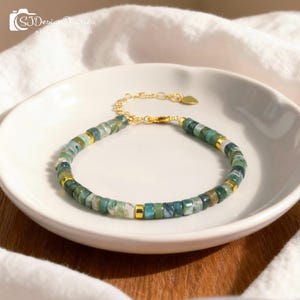 Pode incluir: Uma pulseira de contas verdes e douradas. A pulseira apresenta pequenas contas quadradas em tons de verde e dourado, unidas por uma corrente e um fecho dourados. A pulseira é exibida em um prato branco.