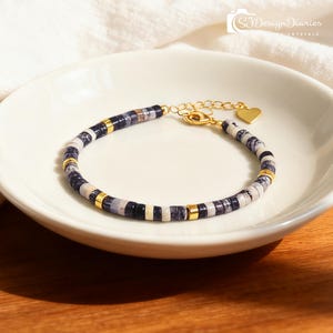 May include: A beaded bracelet with a gold clasp and heart charm. The bracelet features alternating dark blue, white, and gold-colored beads. The bracelet is displayed on a white ceramic plate. The text "SJDesignDiaries CRYSTALS" is in the upper right corner.
