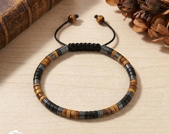 May include: A beaded bracelet featuring alternating segments of brown tiger eye, matte black, and metallic gray disc-shaped beads. The bracelet includes a black cord with an adjustable sliding knot closure, finished with two brown tiger eye beads at the ends.