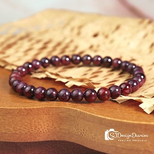 May include: A bracelet made of polished, round, deep red garnet beads. The beads are strung together, forming a complete circle. The bracelet is displayed on a wooden surface with a textured paper background.