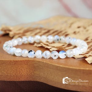 May include: A white moonstone bead bracelet. The round beads have a milky white color with black and blue inclusions. The bracelet is displayed on a wooden surface with a piece of brown paper in the background. The text "SJDesignDiaries Healing Crystals" is in the bottom right corner.