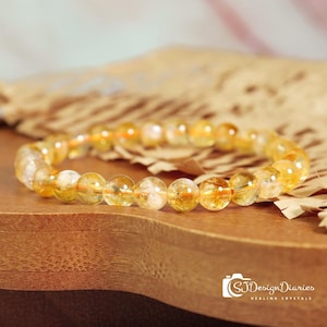 May include: A bracelet made of translucent, golden-yellow citrine gemstone beads. The beads are round and strung together, resting on a wooden surface. The bracelet is a piece of jewelry.