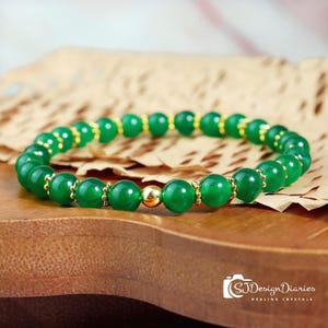 May include: A green beaded bracelet with gold accents. The bracelet features round, polished green beads interspersed with gold-colored spacers. A single, larger gold bead is centered in the design. The bracelet is displayed on a wooden surface.