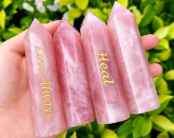 Op de afbeelding: Vier roze rozenkwarts kristallen punten. Twee kristallen hebben de woorden "Love Always" in gouden letters, en twee hebben het woord "Heal". De kristallen worden in een hand gehouden, met een vage groene en witte achtergrond.