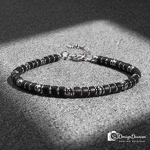 Op de afbeelding: Een zwarte kralenarmband met zilverkleurige metalen accenten. De armband heeft kleine, cilindrische zwarte kralen afgewisseld met zilverkleurige metalen ringen. De sluiting en ketting zijn ook zilver. De armband wordt gepresenteerd op een grijze ondergrond.