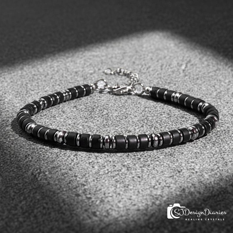 May include: A bracelet featuring alternating matte black cylindrical beads and polished silver-toned metallic spacers. The piece includes a silver-toned lobster claw clasp and an adjustable chain extension. The text SJDesignDiaries is visible.