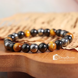 May include: A beaded bracelet featuring a mix of polished black, silver, and tiger's eye gemstones. The bracelet is displayed on a wooden surface, showcasing the natural colors and textures of the stones. The bracelet is a piece of jewelry.