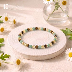 May include: A beaded bracelet featuring a repeating pattern of round, polished tiger eye, citrine, and green aventurine gemstones. The beads are strung on a flexible cord, creating a circular shape with a smooth, glossy finish on each stone.