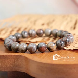 May include: A beaded bracelet featuring round, polished stones in shades of gray, brown, and red. The bracelet is displayed on a wooden surface with a piece of parchment paper. The bracelet is a piece of jewelry.