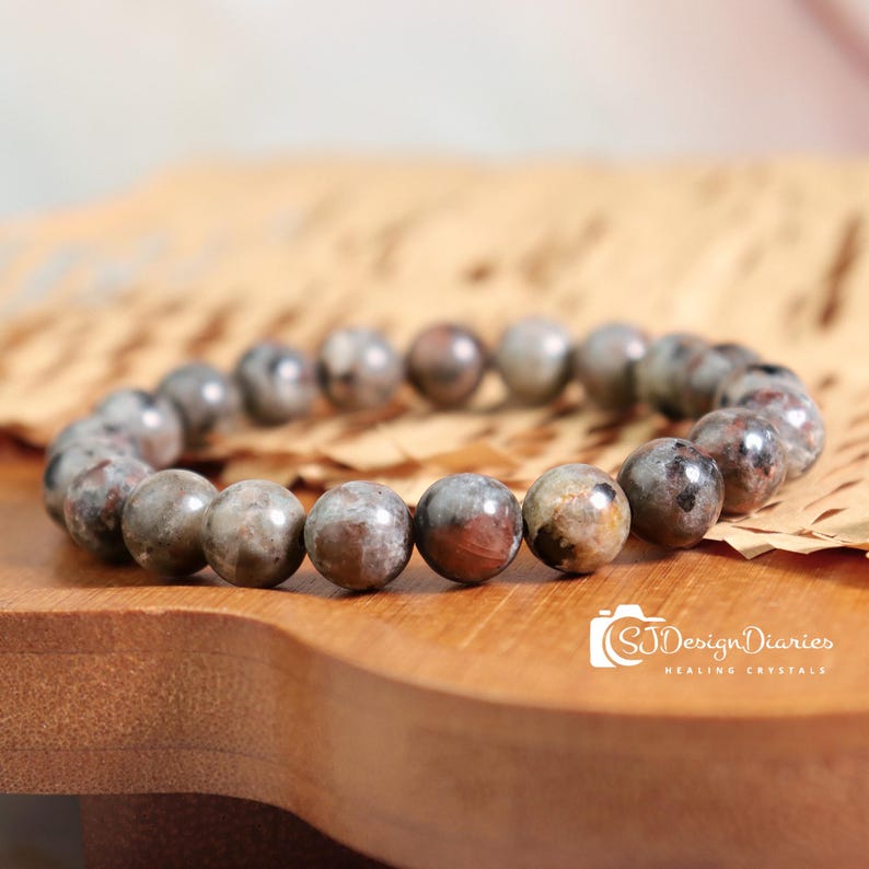 May include: A beaded bracelet featuring round, polished stones in shades of gray, brown, and red. The bracelet is displayed on a wooden surface with a piece of parchment paper. The bracelet is a piece of jewelry.