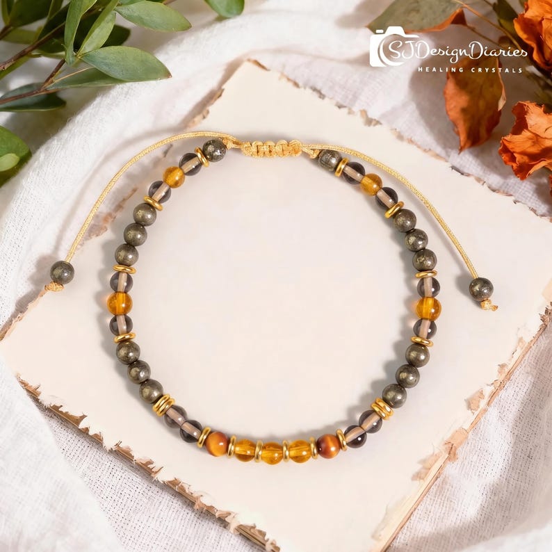 K&ouml;nnte beinhalten: Ein Perlenarmband mit abwechselnden metallischen Pyritkugeln, durchscheinenden Rauchquarzperlen und leuchtend bernsteinfarbenen Steinen. Das Design enth&auml;lt kleine goldfarbene Zwischenelemente und einen beigen, verstellbaren Kordelverschluss.