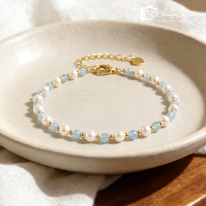 May include: Elegant gold bracelet with alternating white pearls and light blue beads. The bracelet has small gold beads and a gold clasp. The bracelet is presented on a cream-coloured plate.