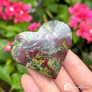 May include: A heart-shaped stone with a mix of red, green, and brown hues. The stone is held in a hand, with a blurred background of pink flowers and green foliage. The stone's surface has a natural, textured appearance.