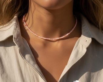 Könnte beinhalten: Eine zarte, rosa Perlenkette liegt auf dem Hals einer Person. Die Halskette besteht aus kleinen, gleichmäßig verteilten Perlen, die einen subtilen, eleganten Look erzeugen. Die Person trägt ein helles Hemd mit Knöpfen.