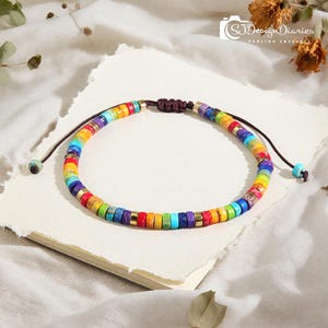 May include: A rainbow-colored beaded bracelet with a brown adjustable cord. The bracelet features a variety of colors, including red, orange, yellow, green, blue, and purple. The beads are separated by small gold-colored spacers. The bracelet is on a white surface.