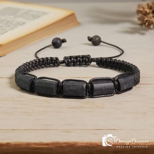 May include: Black macrame bracelet featuring rectangular black stones. The bracelet has an adjustable sliding knot closure. The background includes a book and a wooden surface. The bracelet is a piece of jewelry.